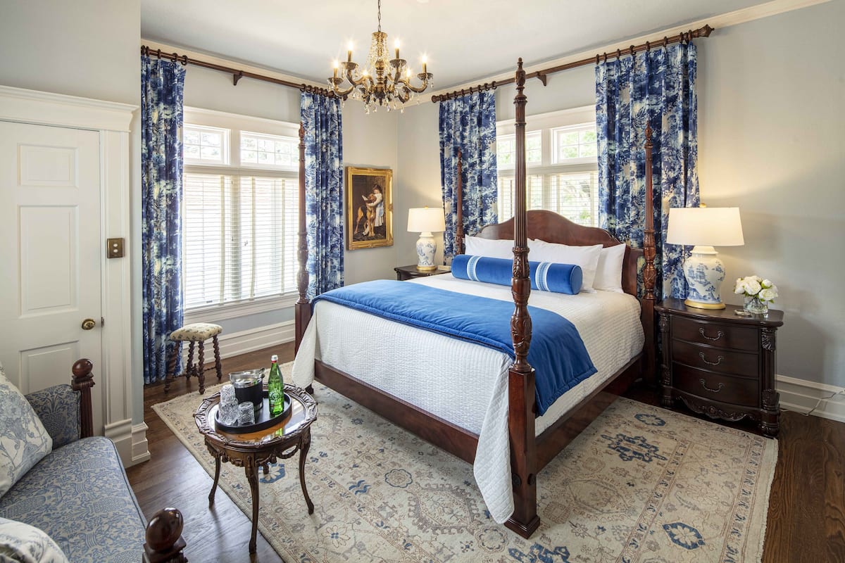 A four-poster bed with a blue and white bedspread, a chandelier, and a nightstand with a lamp.