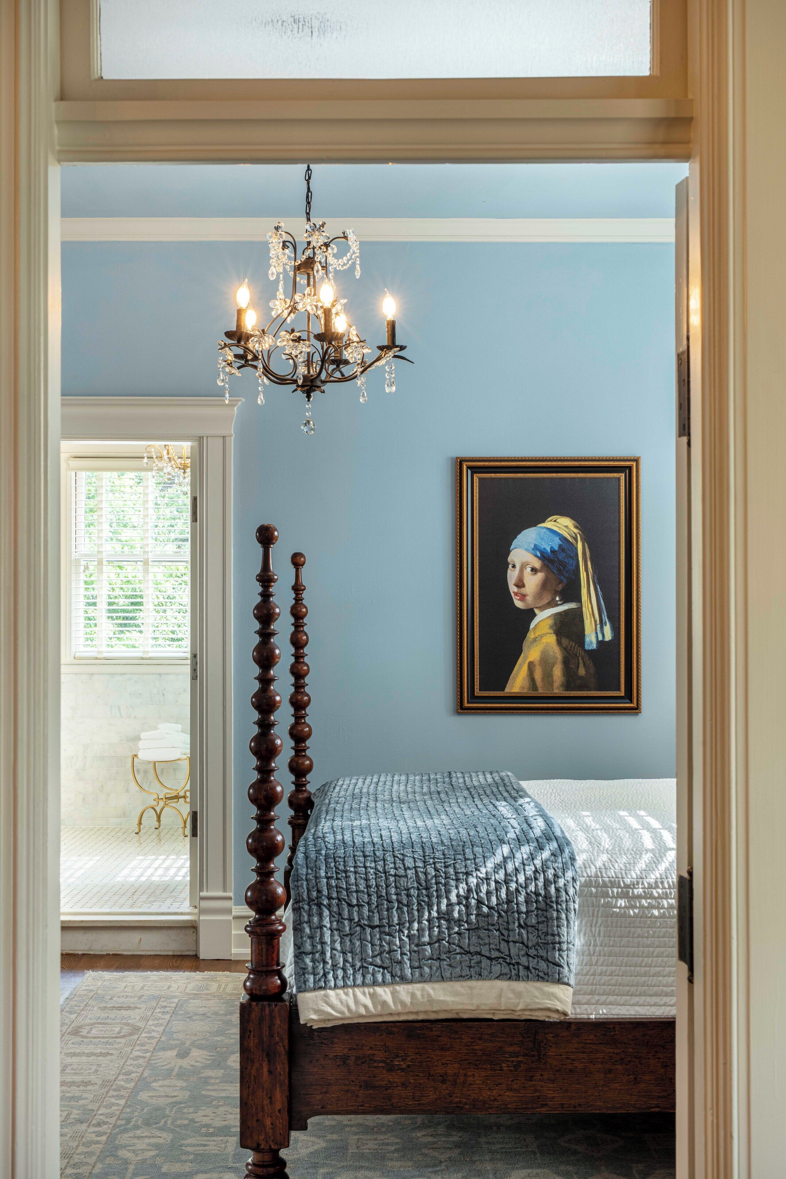 A bedroom with a four-poster bed, a chandelier, a framed artwork on the wall, and a view of the outside through a door.