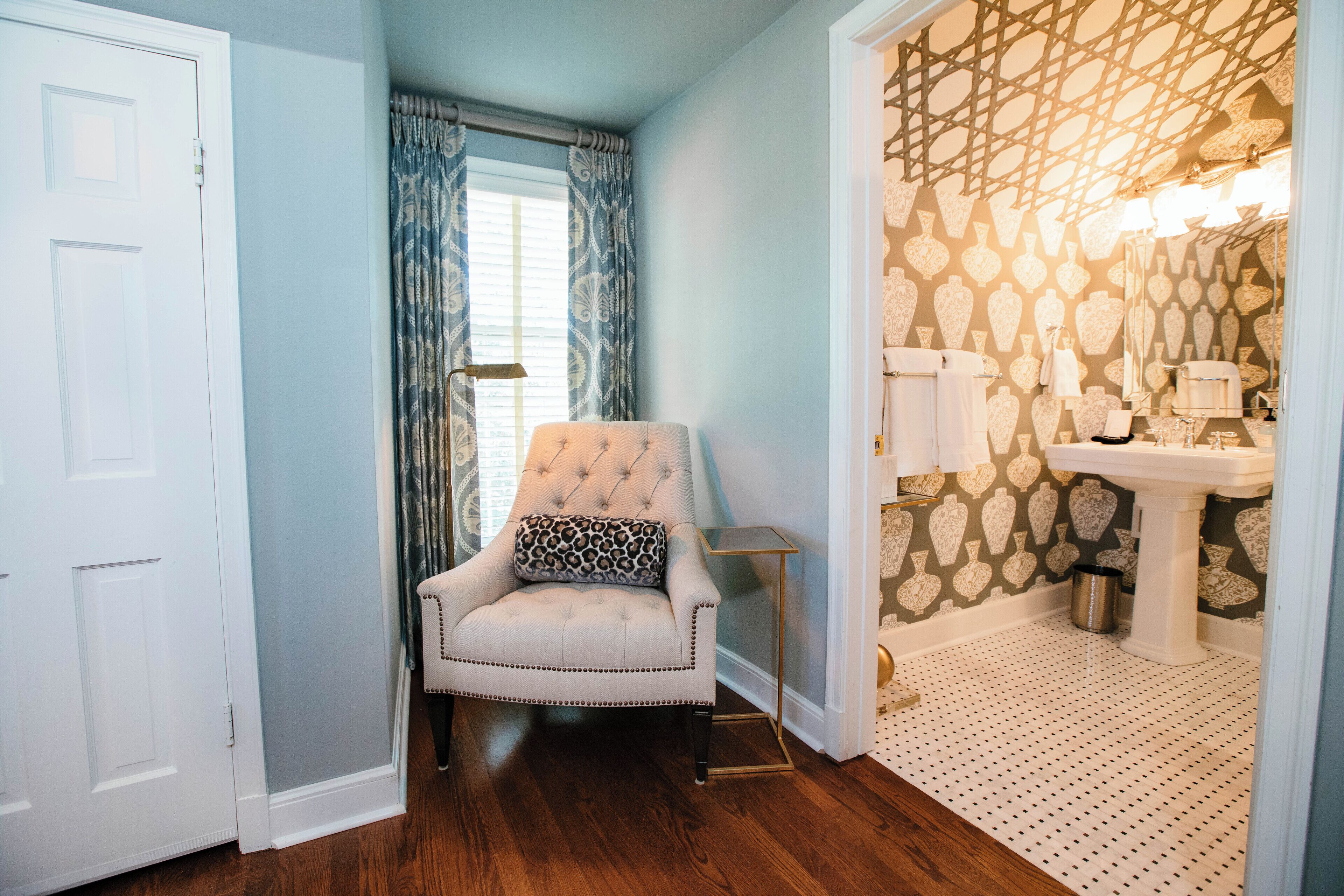 A room with a tufted armchair, a patterned wallpaper, a white door, and a bathroom with a sink and mirror.