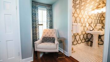 A room with a tufted armchair, a patterned wallpaper, a white door, and a bathroom with a sink and mirror.