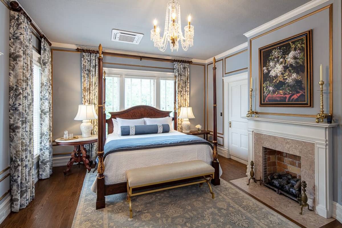 A four-poster bed with a wooden headboard, two bedside tables with lamps, a bench, a fireplace, and a framed painting on the wall.