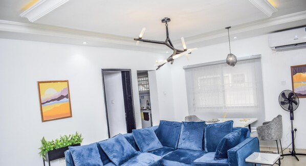 A modern living room with a blue sectional sofa, a small table, a fan, and a ceiling light fixture.