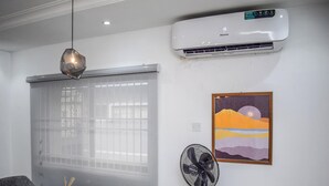 Un salon moderne avec une table à manger, un fauteuil gris, un ventilateur sur pied et un appareil de climatisation.