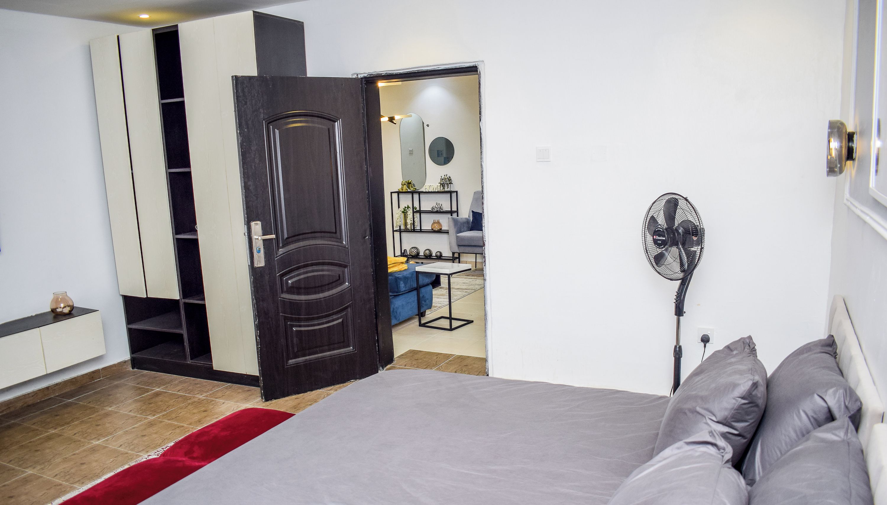 A bedroom with a bed, a red throw, a wardrobe, a door, a fan, and a view into another room with a chair and a shelf.