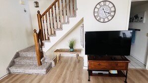 A living room with a wooden staircase, a flat-screen TV on a wooden stand, and a wall clock.