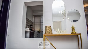 A room with a white wall, a window with a view of a kitchen, a round mirror, a gold-colored table with a lamp, and a gold statue.