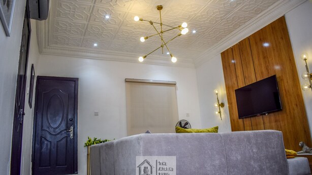 A modern living room with a grey sofa, a wooden accent wall, a mounted TV, and a decorative ceiling light fixture.