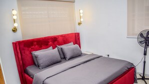 A bed with a red headboard and grey bedding, two wall-mounted light fixtures, a standing fan, and a window with blinds.