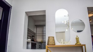 A room with a white wall, a window with a view of a kitchen, a round mirror, a gold-colored table with a lamp, and a gold statue.