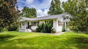 Une maison de plain-pied avec un porche à l’avant, entourée d’une pelouse bien entretenue et d’arbres.