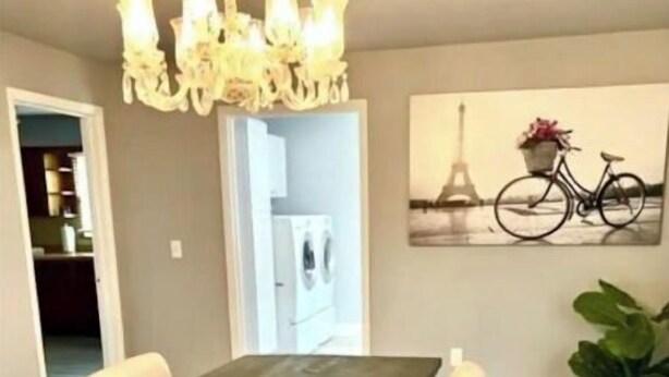 Un coin repas avec une table en bois, des chaises à revêtement blanc, un lustre, un tableau représentant un vélo accroché au mur et une plante en pot.