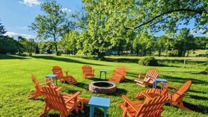 Un ensemble de sièges d’extérieur disposés en cercle autour d’un foyer central, le tout étant entouré d’une pelouse et d’arbres.