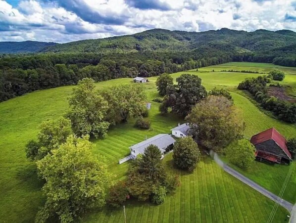 Ländliche Landschaft mit einem Haus, umgeben von üppigen grünen Feldern und Bäumen, mit Hügeln im Hintergrund.