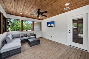 A covered outdoor living area with a wooden ceiling, ceiling fan, and large windows offering a view of greenery.
