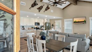 A spacious kitchen with white cabinets, a dining area with a wooden table and white chairs, and a gray sofa in the living area. - Farmhouse Charm - Waco/Magnolia/Baylor (Waco)