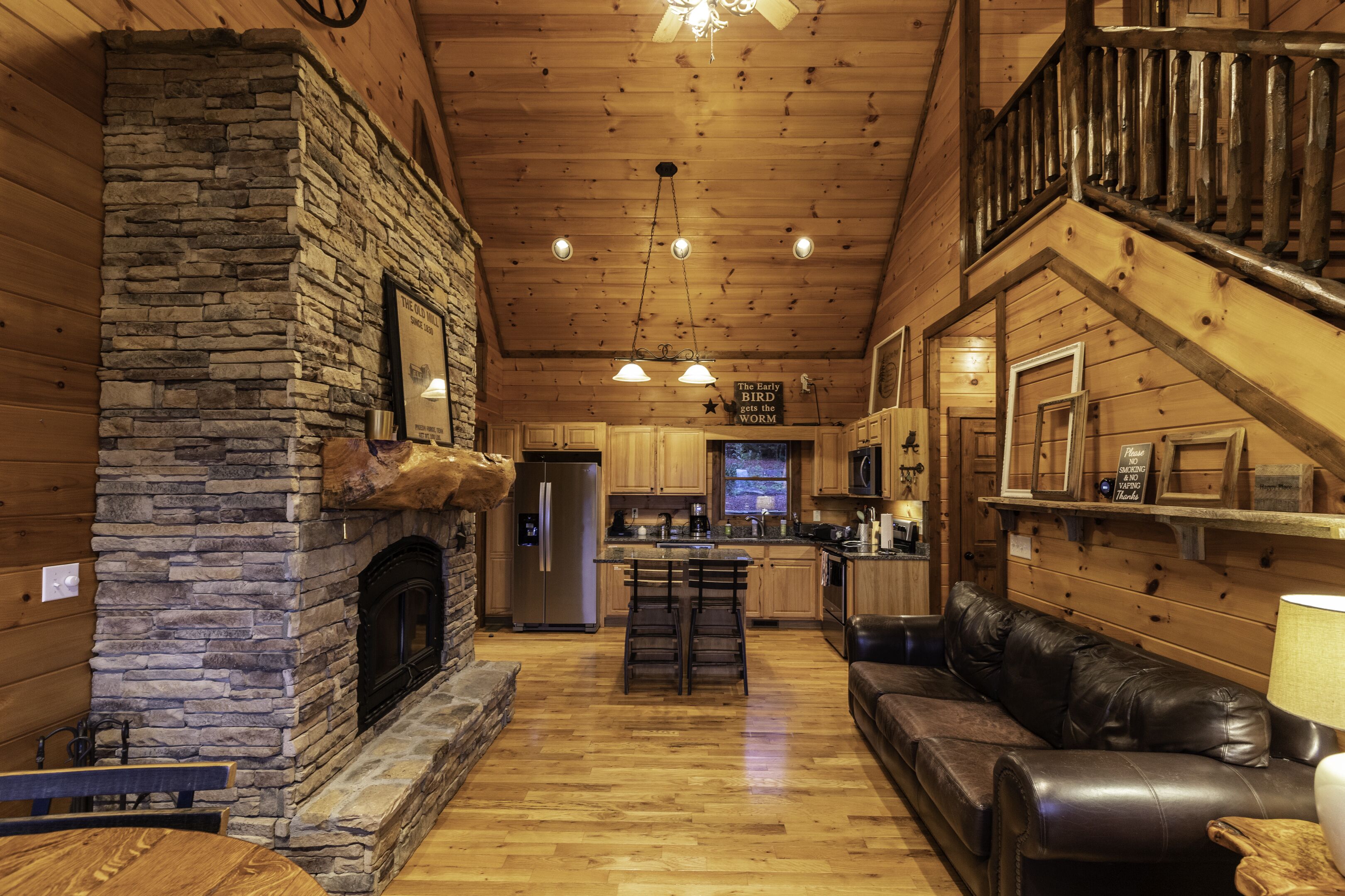 A cozy cabin interior with a stone fireplace, wooden ceiling, a kitchen area, and a leather sofa.