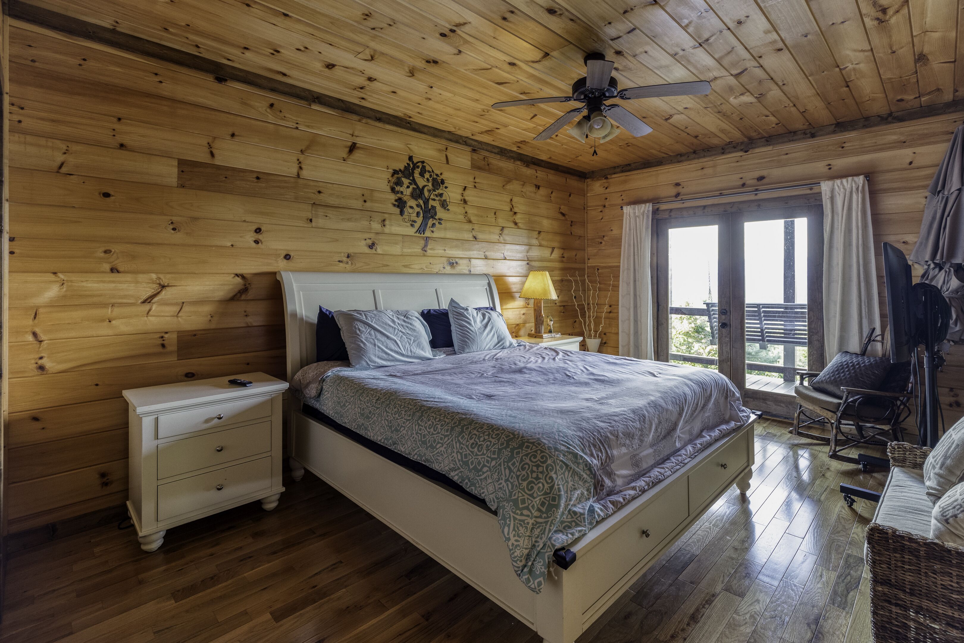 A wooden-paneled bedroom with a bed, nightstands, a ceiling fan, and a view through a window.