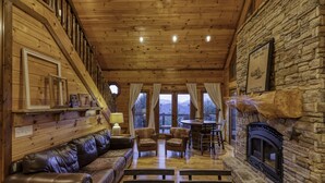 Interior acogedor de una cabaña con chimenea de piedra, paredes de madera, un sofá de cuero y un comedor con vistas a las montañas.
