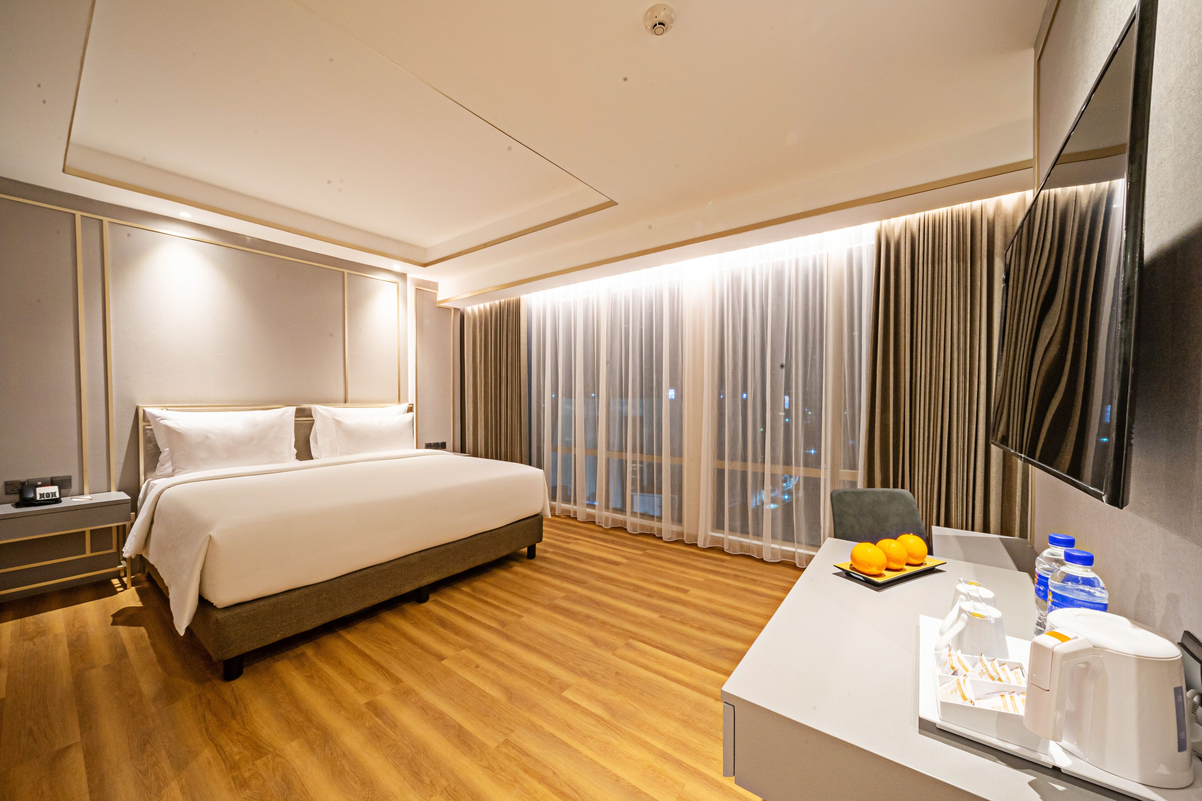 Habitación de hotel con una cama grande, un escritorio con cafetera y vistas a la ciudad a través de cortinas translúcidas.