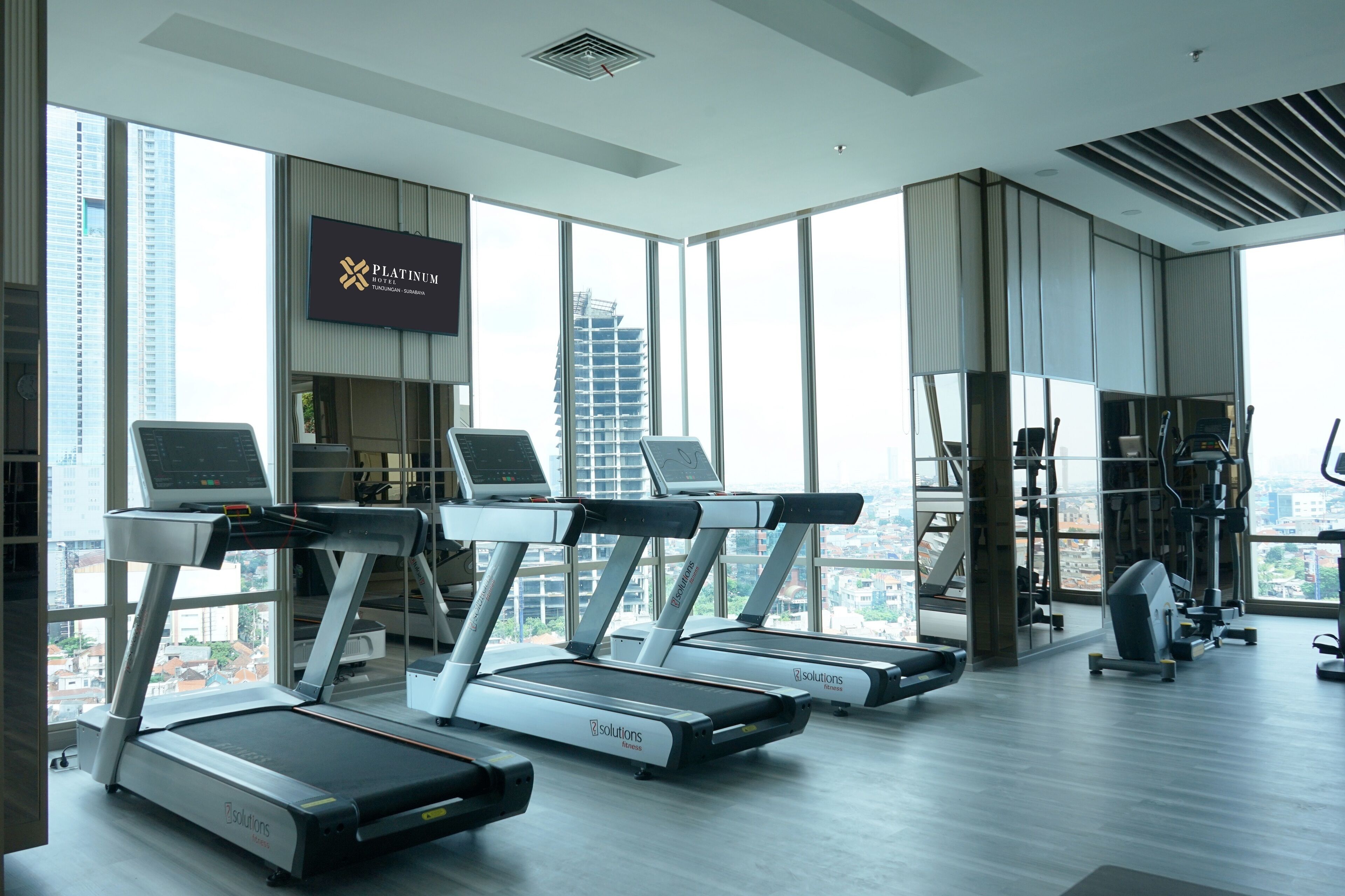 Une salle de sport moderne équipée de tapis de course, d’appareils d’exercice et de grandes fenêtres offrant une vue sur la ville.