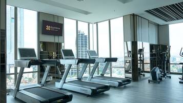 Une salle de sport moderne équipée de tapis de course, d’appareils d’exercice et de grandes fenêtres offrant une vue sur la ville.