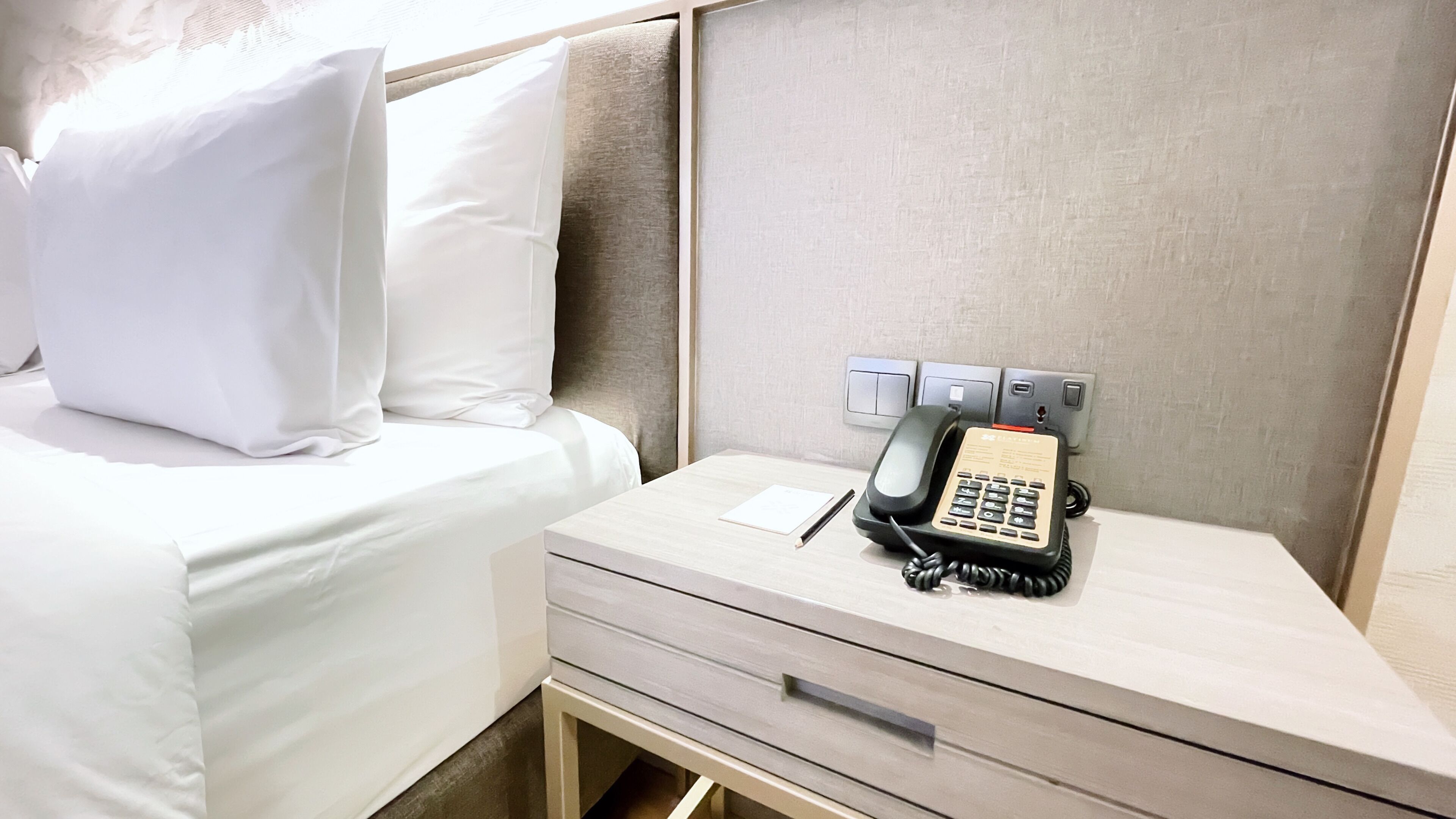 Ein Hotelzimmer mit einem Bett, einem Nachttisch mit Telefon, Notizblock und Stift.