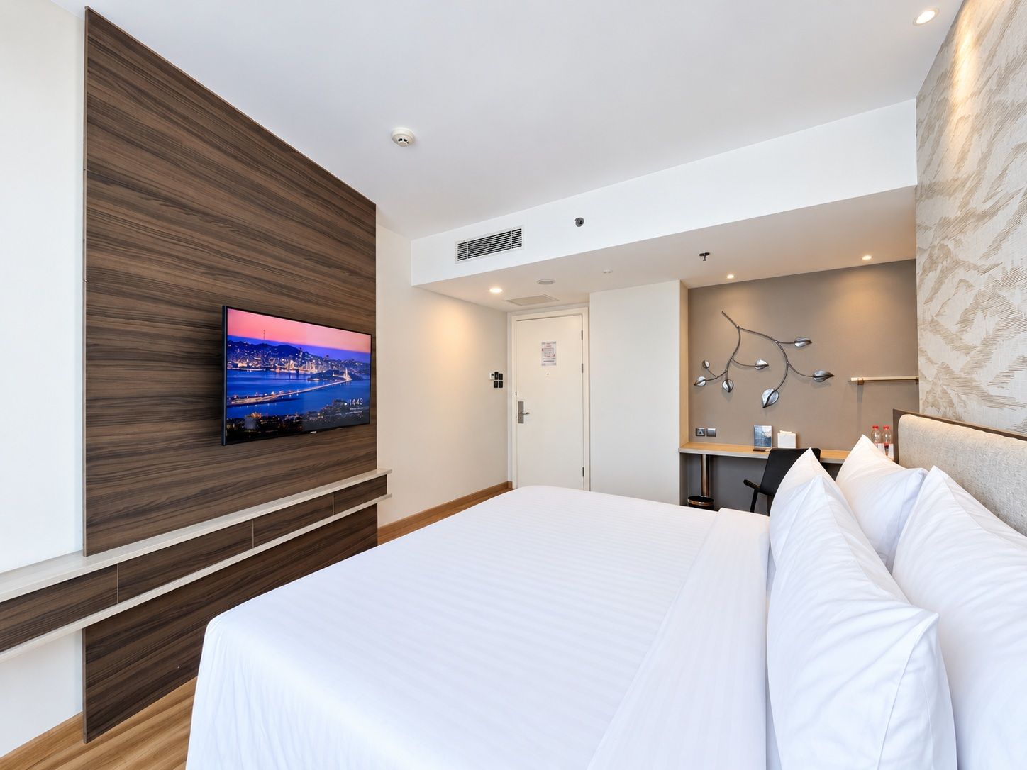 Hotellihuone, jossa on suuri sänky, seinälle asennettu televisio, josta näkyy kaupunkimaisema, työpöytä tuolilla sekä seinään kiinnitetty valaisin.