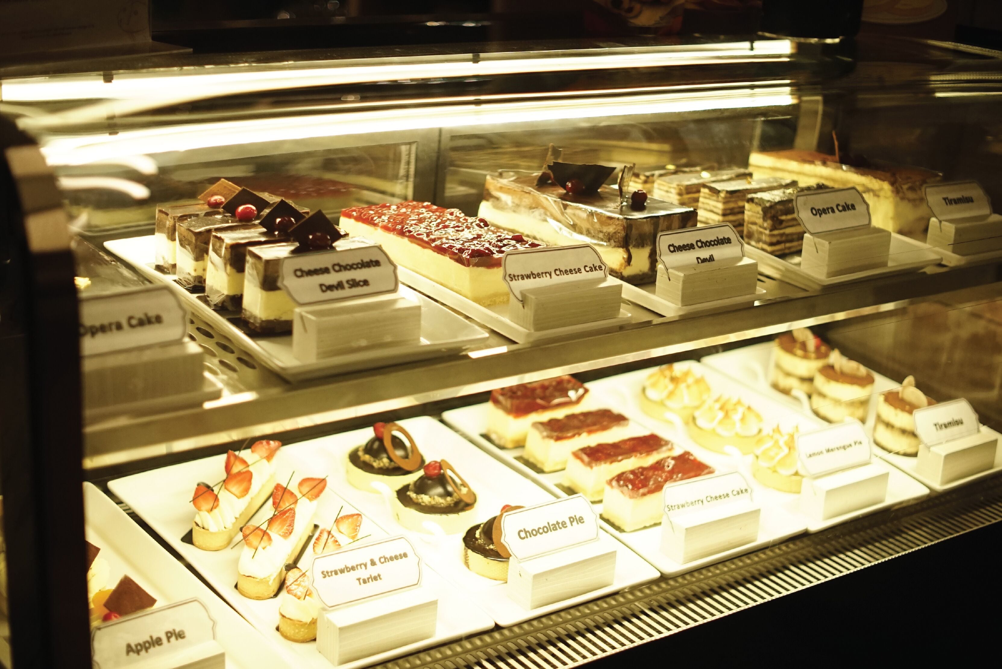 Una vitrina con diversos postres, incluyendo etiquetas como 'Opera Cake', 'Strawberry Cheese Cake' y 'Chocolate Pie'.