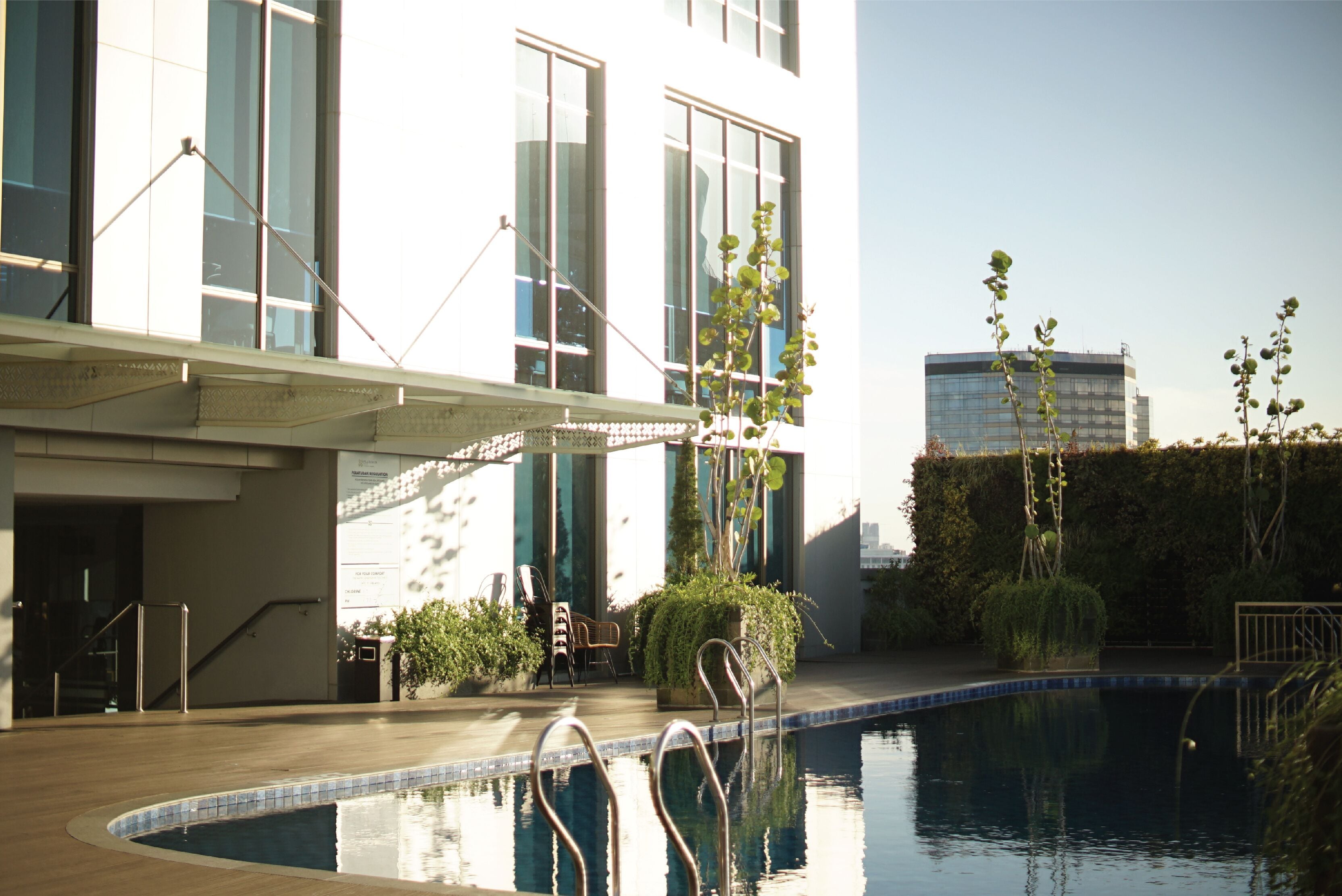 Una piscina con escaleras metálicas, rodeada por una terraza de madera, y contigua a un edificio con amplias ventanas y una zona de estar exterior cubierta.