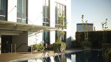 Una piscina con escaleras metálicas, rodeada por una terraza de madera, y contigua a un edificio con amplias ventanas y una zona de estar exterior cubierta.