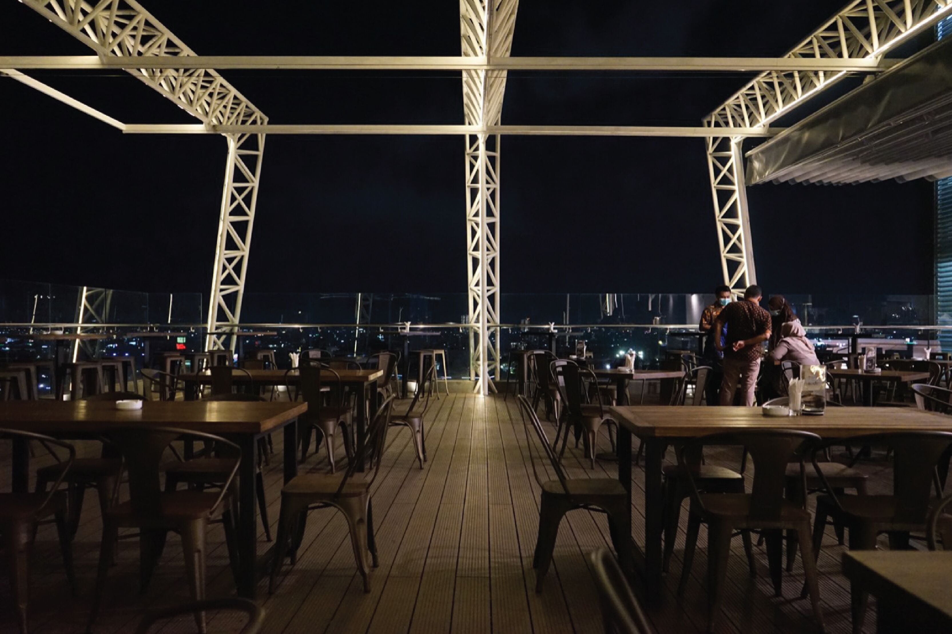 An outdoor dining area with tables and chairs, a view of the cityscape, and a truss structure overhead.