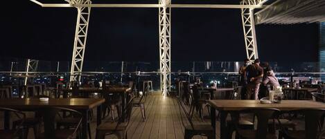 An outdoor dining area with tables and chairs, a view of the cityscape, and a truss structure overhead.