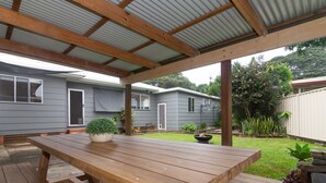 A covered patio area with a wooden table and benches, surrounded by a garden and a house with a grey exterior. - Charming 3 bedroom, modern, waterfront home (North Haven)