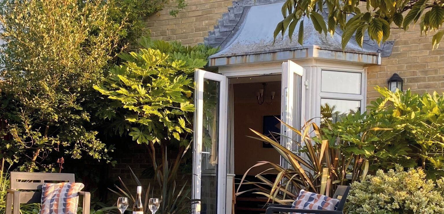 A cozy outdoor seating area with a wooden bench and a small table, surrounded by lush greenery and a glass enclosure.