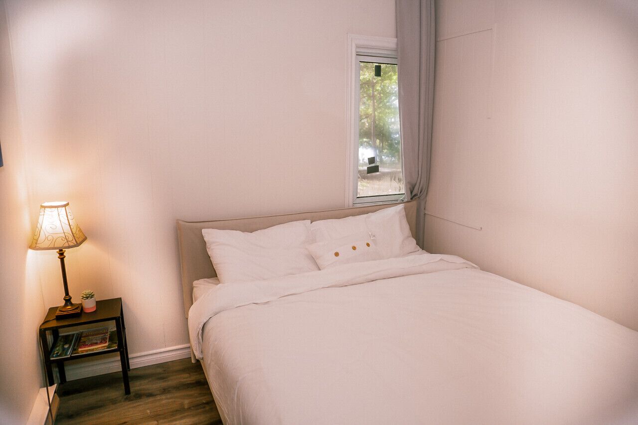 Un dormitorio con cama, mesita de noche con lámpara, estante pequeño y ventana con cortinas.