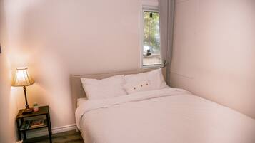 Un dormitorio con cama, mesita de noche con lámpara, estante pequeño y ventana con cortinas.