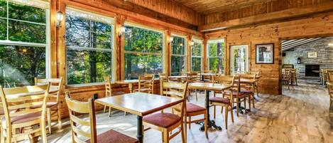 A spacious wooden interior with large windows, wooden tables and chairs, and a stone fireplace.