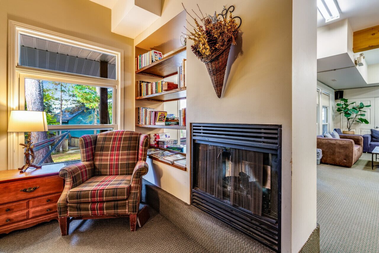 Una sala de estar acogedora con un sillón de cuadros, una chimenea, una estantería y un ventanal con vistas a un jardín trasero.