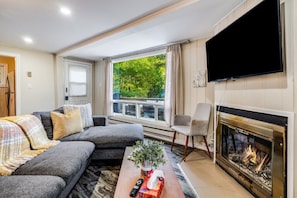 Una sala de estar con un sofá, una chimenea, un televisor y un ventanal con vistas a árboles.