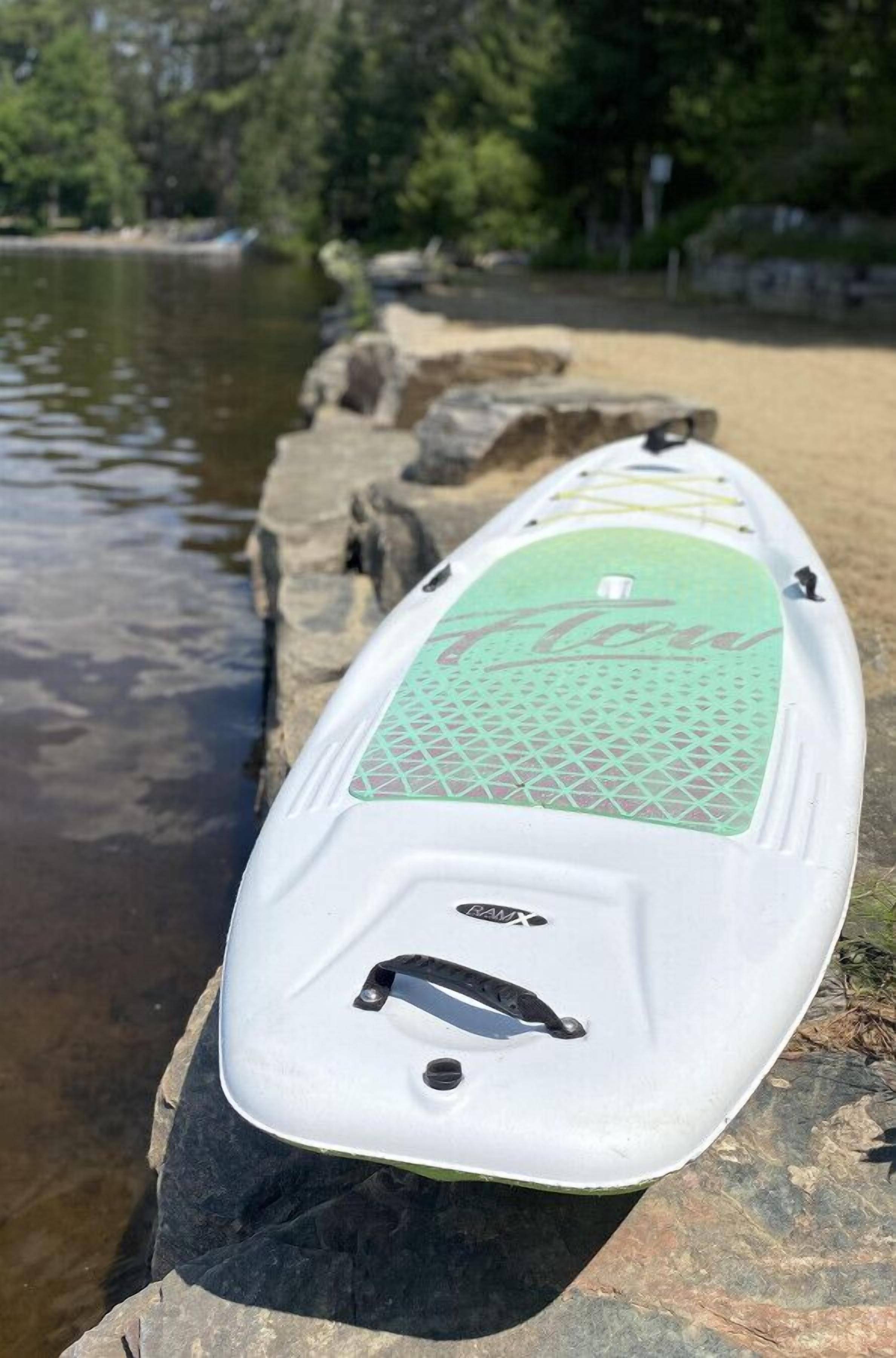 Una tabla de remo blanca con superficie texturizada verde y la palabra "KAYAK" sobre rocas junto a un cuerpo de agua.