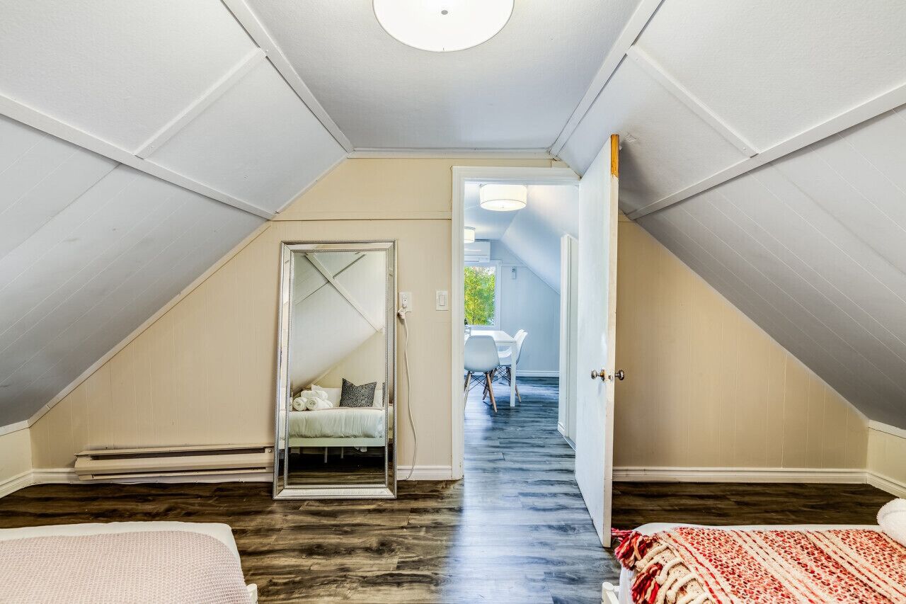 Une petite chambre à coucher de style mansardé, avec un plafond en pente, un lit recouvert d’une courtepointe à motifs rouges et blancs, et une porte visible donnant sur un couloir.