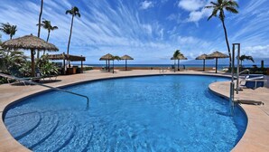 A swimming pool with a curved edge, surrounded by lounge chairs and thatched umbrellas, with a view of the ocean. - Royal Kahana 301 Remodeled OceanFront 2BD w Ocean Views Pool (Lahaina)