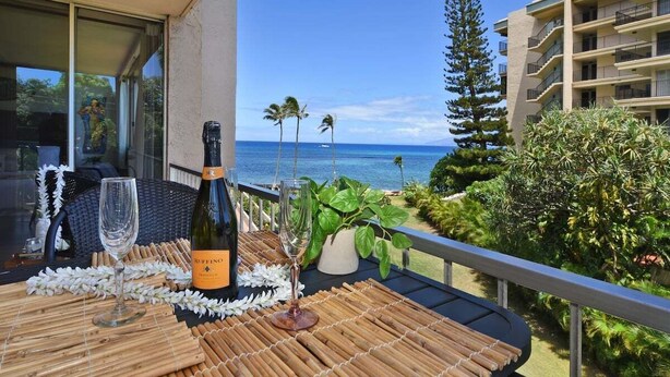 A balcony with a table set for two, featuring a bottle of wine and two glasses. The balcony overlooks a tropical landscape with palm trees and a view of the ocean. - Royal Kahana 301 Remodeled OceanFront 2BD w Ocean Views Pool (Lahaina)