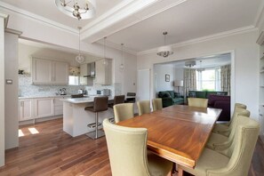 Un vaste espace de salle à manger avec une grande table en bois et six chaises, une cuisine dotée de meubles blancs et un salon comprenant un canapé et une table basse.