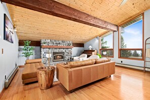 Amplia sala de estar con chimenea de piedra, vigas de techo de madera y grandes ventanas que ofrecen vistas al exterior.