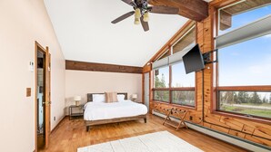 Un dormitorio con piso de madera, una cama con ropa de cama blanca, un ventilador de techo, un televisor montado en la pared y ventanas amplias con vistas al exterior.