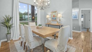 Un espace repas avec une table en bois, des chaises blanches, un lustre et une vue sur l’extérieur.