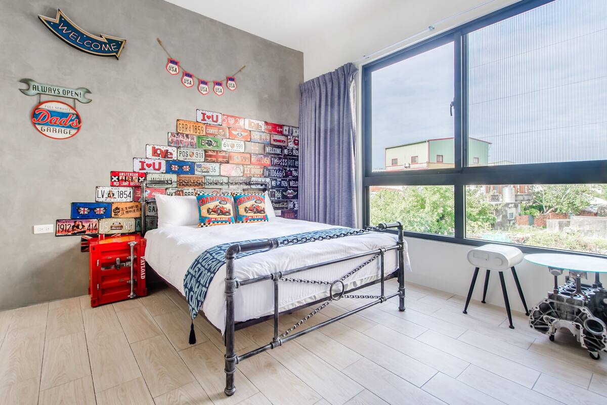 Une chambre à coucher avec un lit en métal, une fenêtre donnant sur un paysage agréable et un mur décoré de plaques d’immatriculation anciennes.