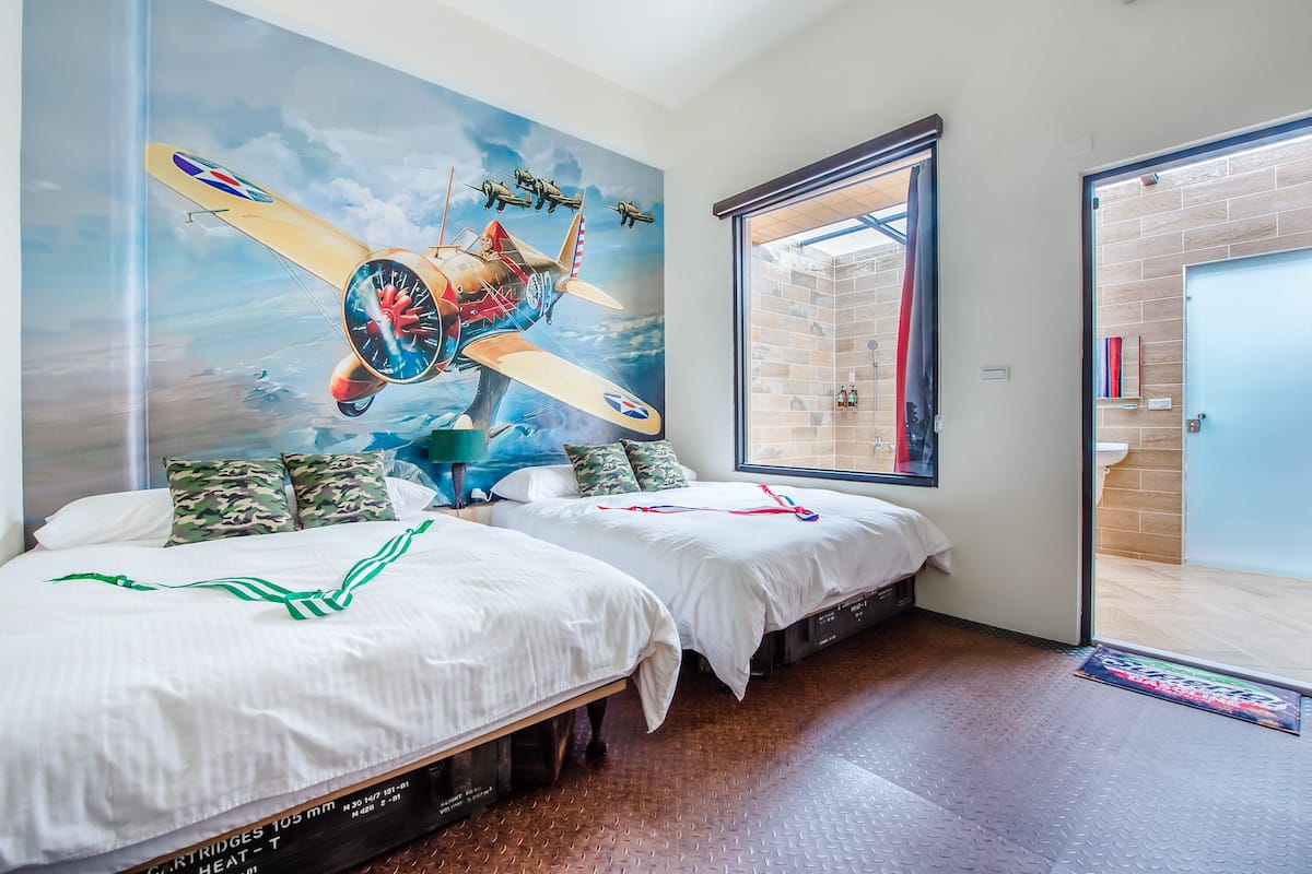 Une chambre d’hôtel avec deux lits, une fresque représentant des avions, une grande fenêtre et une salle de bain visible à travers une porte ouverte.