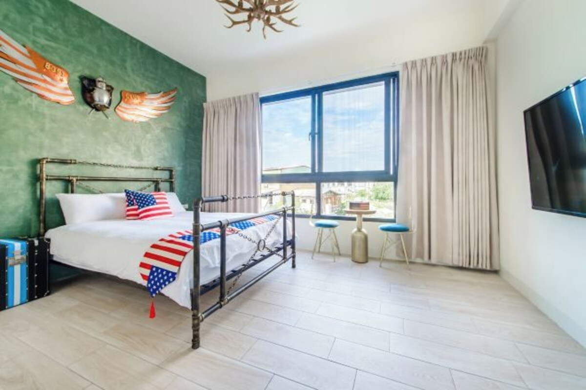 A bedroom with a metal bed frame, a large window with curtains, a TV mounted on the wall, and a small table with chairs.